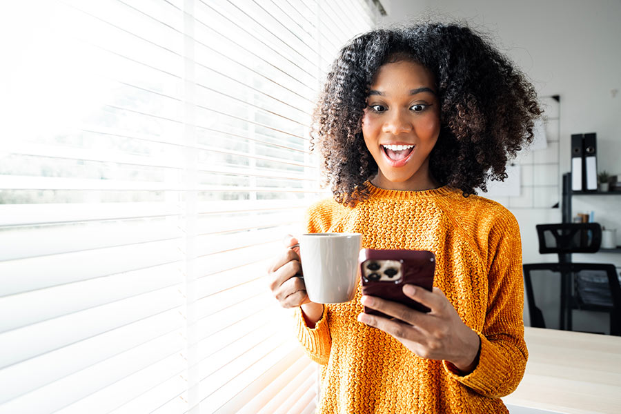 Photo of a young woman holding a cup of coffee and looking at her phone by a window