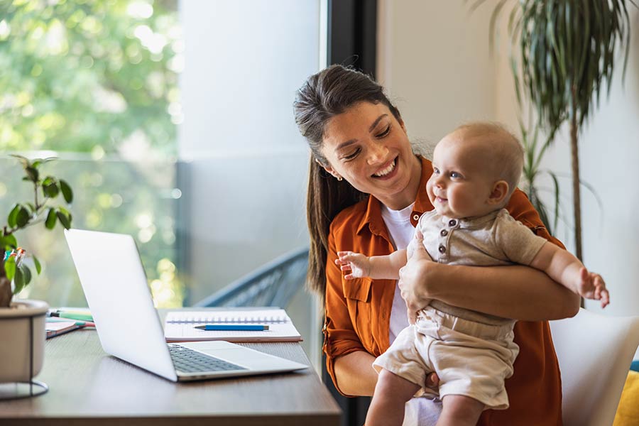 Photo of a smiling woman holding a happy baby while sitting at a table with a laptop